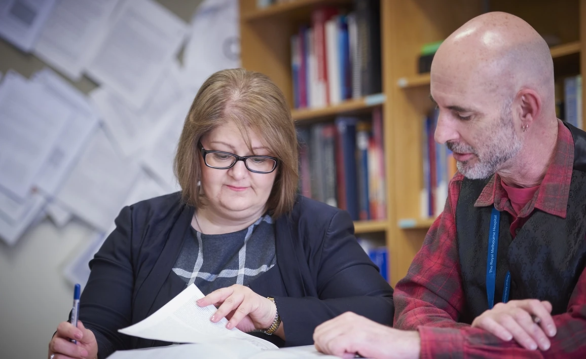 Two professionals review documents together in a research or academic setting, surrounded by bookshelves and pinned papers on the wall.