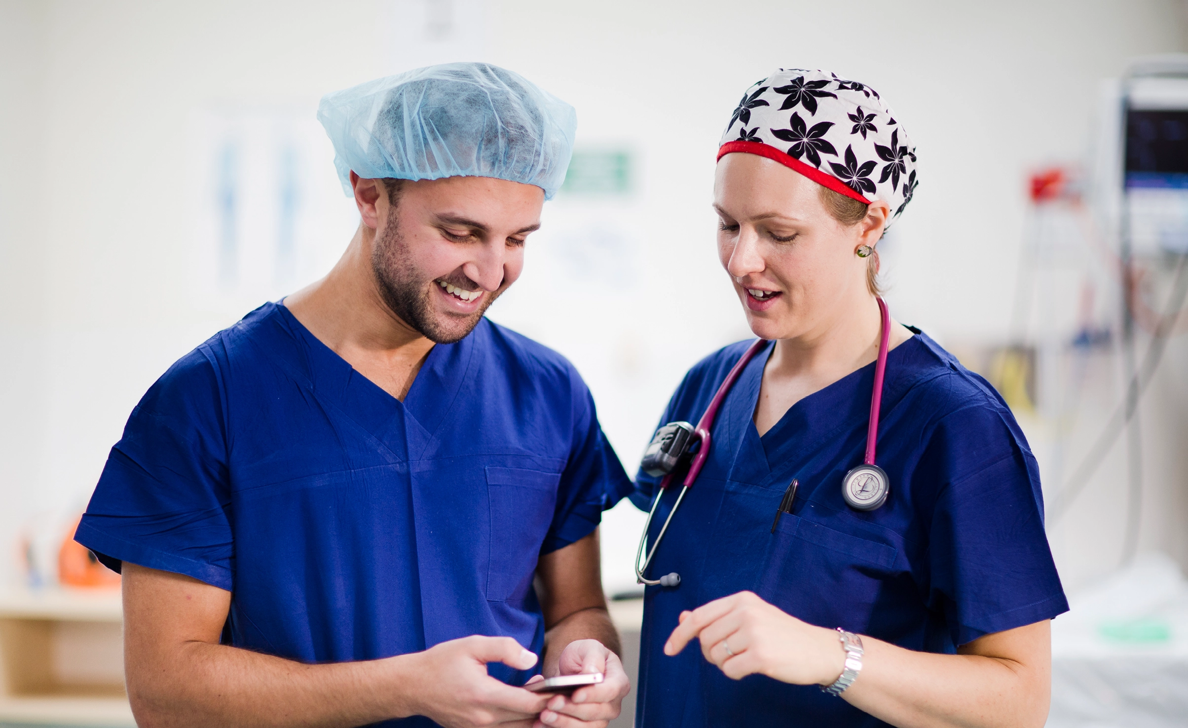 Two people in blue scrubs smiling and looking down at phone