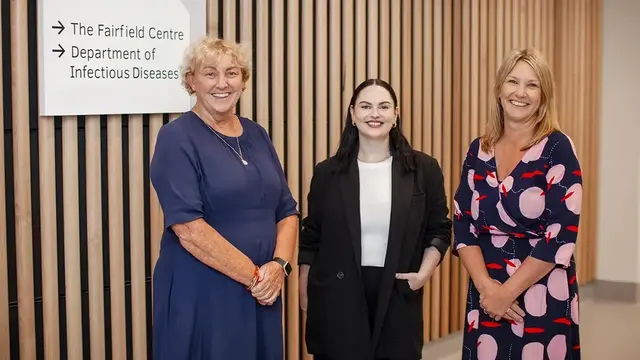Three professionally dressed trial coordinators stand together, smiling, in front of a sign for "The Fairfield Centre Department of Infectious Diseases," against a modern wooden panel backdrop.