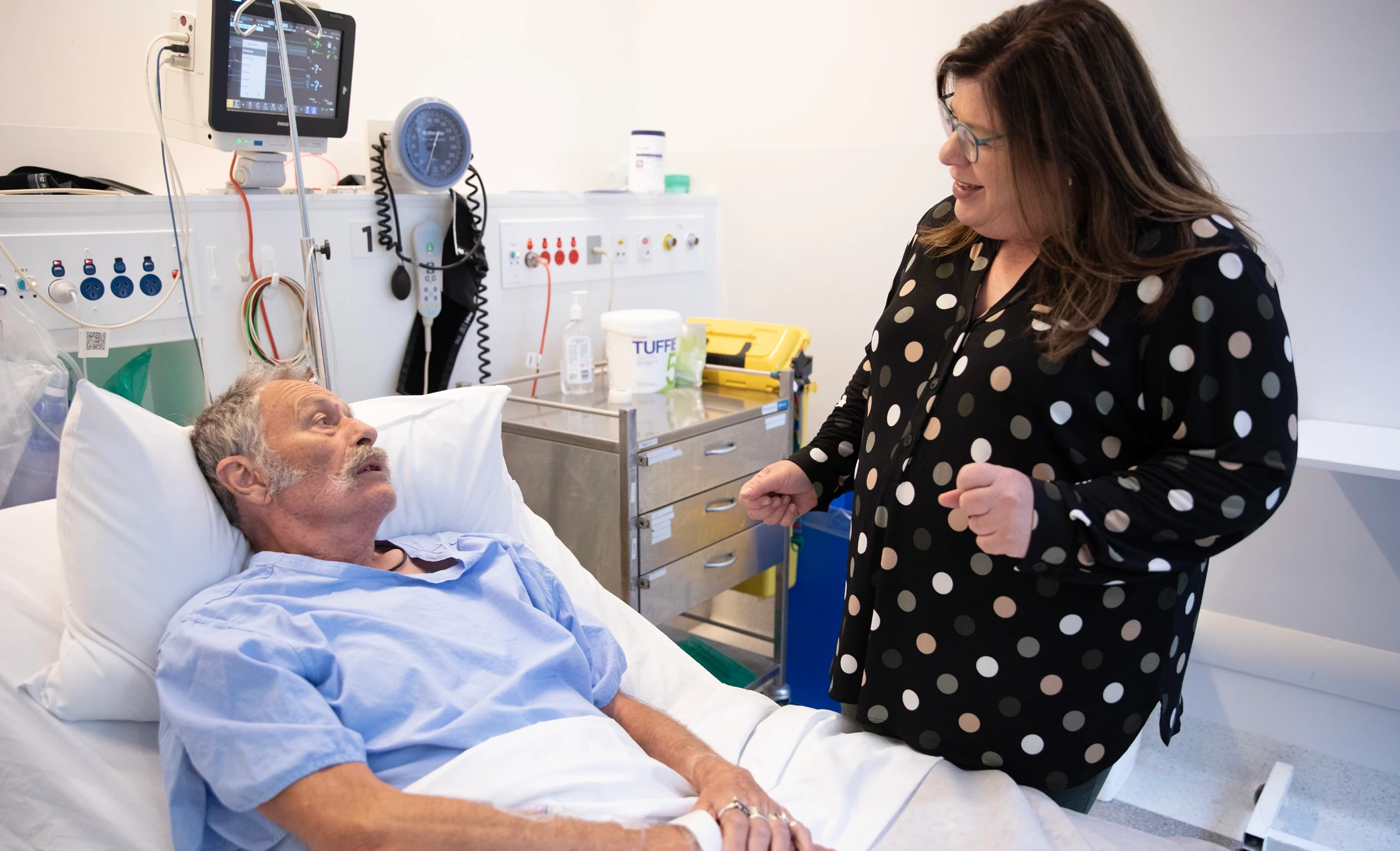 An older patient lies in a bed with a doctor discussing their treatment