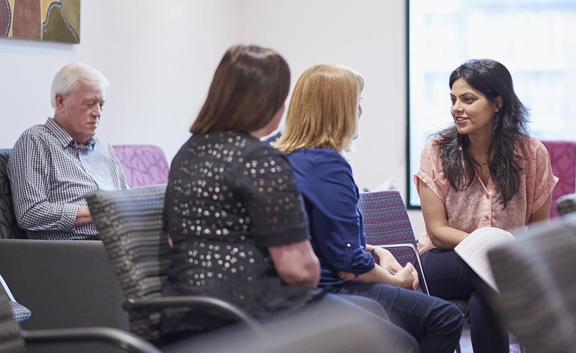 A group of individuals engage in a discussion in a waiting area, with a research coordinator provides informed consent, while another person sits nearby reading quietly.