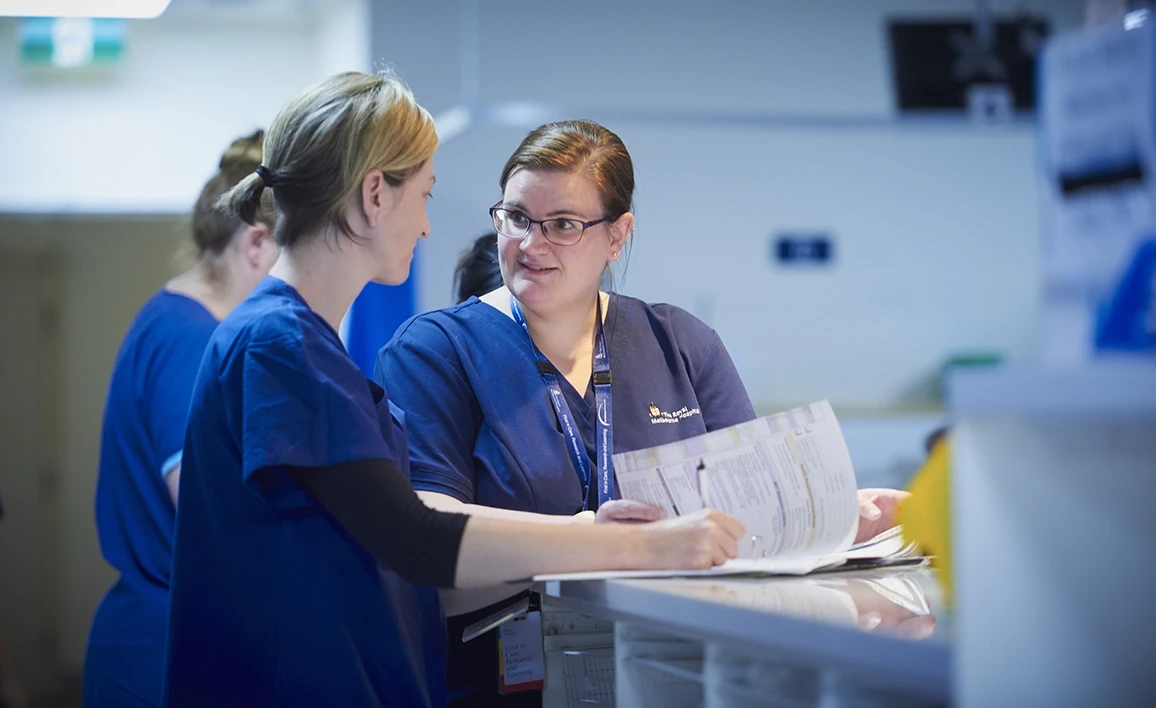 Two healthcare professionals in navy scrubs discuss patient documentation at a hospital workstation, with a busy clinical environment in the background.