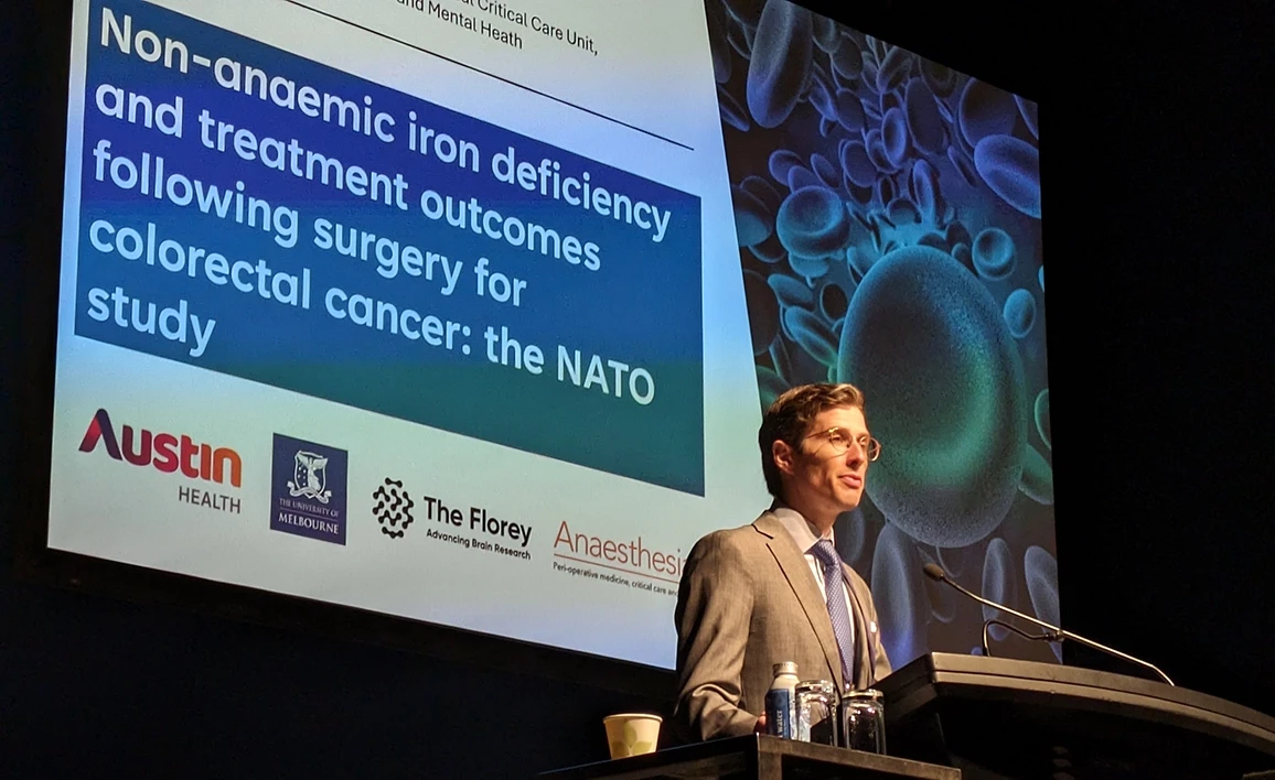 An anaesthetist in a suit stands at a podium, delivering a talk on the NATO study about non-anaemic iron deficiency and treatment outcomes after colorectal cancer surgery, with a large presentation screen behind him.