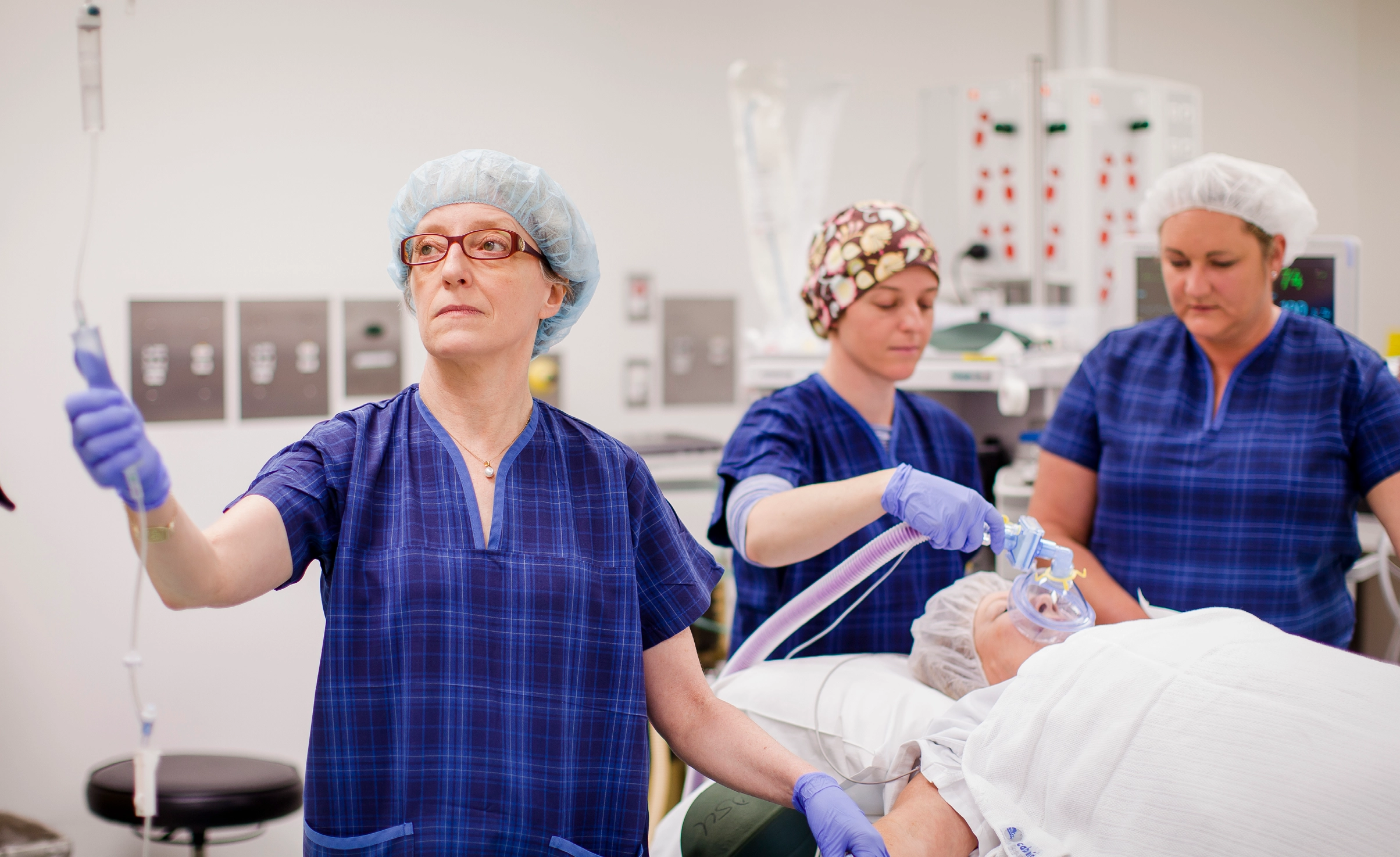 Person in scrubs checking IV fluids. Two people in scrubs in the background check on patient with anaesthesia mask on