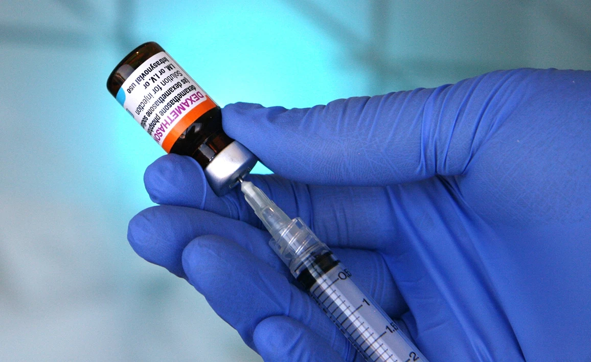 A gloved healthcare professional draws Dexamethasone from a vial into a syringe against a cool-toned, clinical background.