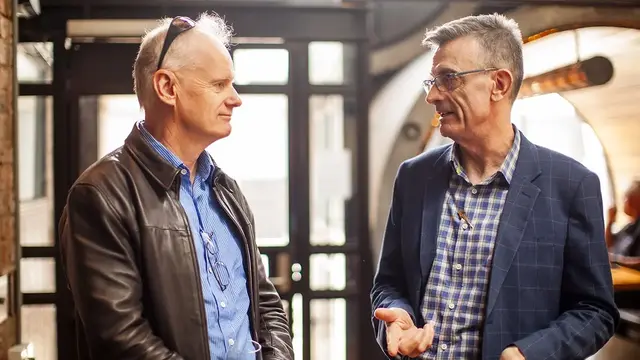 Two professionally dressed men engaged in a conversation in a well-lit indoor setting.