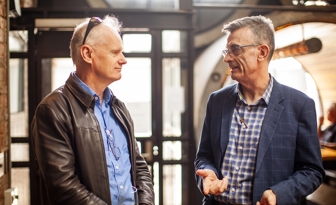 Two professionally dressed men engaged in a conversation in a well-lit indoor setting.