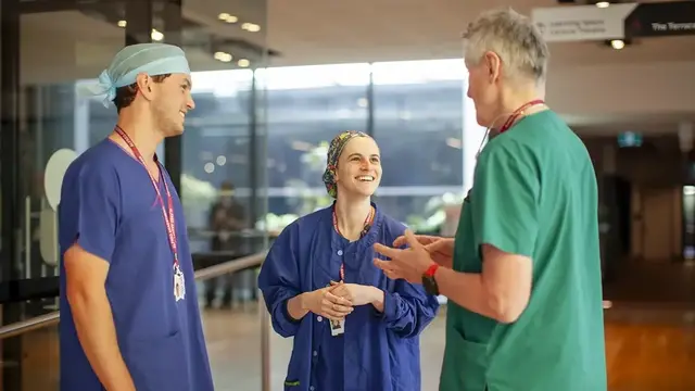 The image shows three healthcare professionals in scrubs having a lively and friendly conversation in a hospital or clinical setting. 