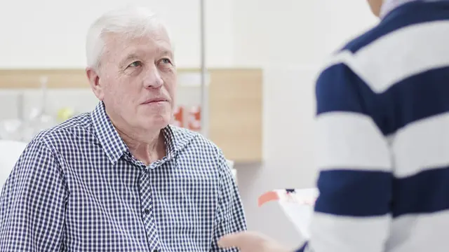 An older patient in a checkered shirt listens attentively to a discussion with an anaesthetist holding a document in a clinical setting.