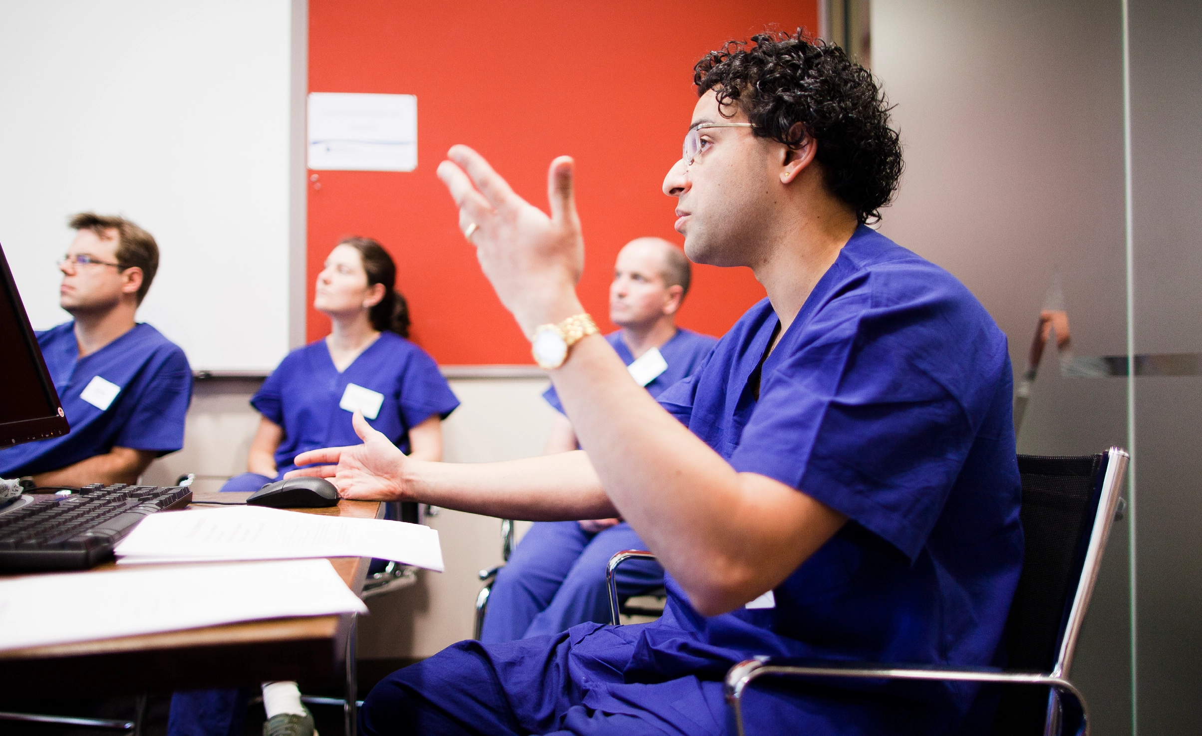 Person in scrubs in front of a computer talking with three people in scrubs in the background