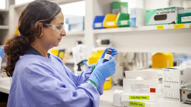 A researcher in protective gear, including gloves and safety glasses, examines a vial in a laboratory setting, with labeled study materials in the background.