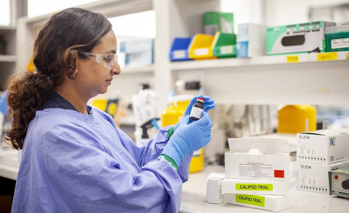 A researcher in protective gear, including gloves and safety glasses, examines a vial in a laboratory setting, with labeled study materials in the background.