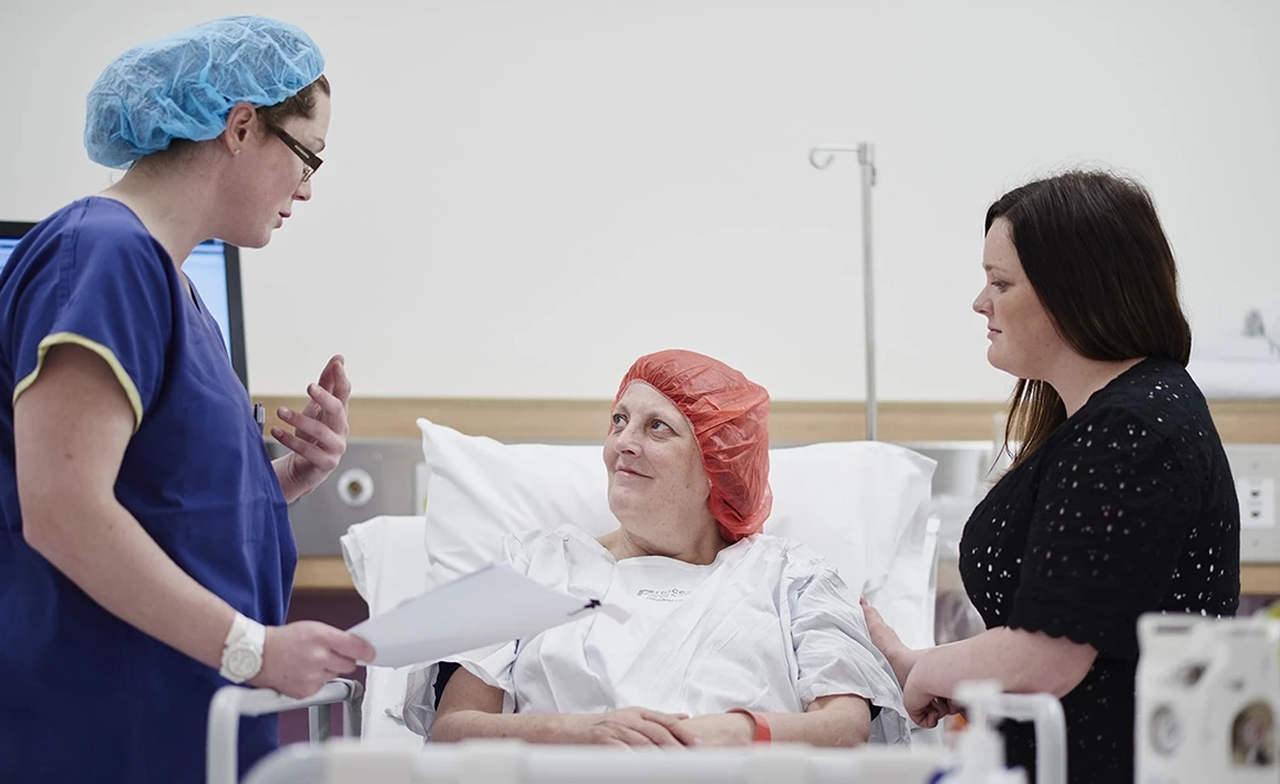 An anaesthetist in scrubs speaks with a patient in a hospital bed, who is accompanied by a visitor, in a pre-surgery setting.