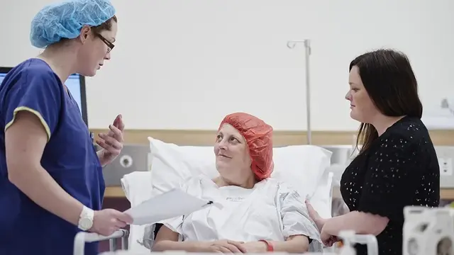An anaesthetist in scrubs speaks with a patient in a hospital bed, who is accompanied by a visitor, in a pre-surgery setting.