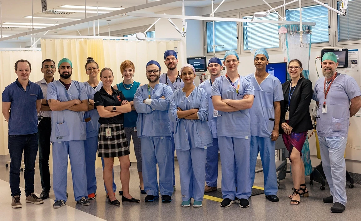 A diverse team of healthcare professionals, including surgeons, nurses, and administrators, pose together in a hospital setting, dressed in scrubs and professional attire.