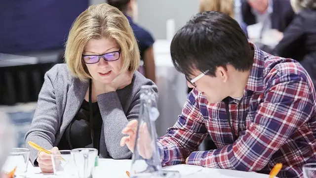 Two professionals collaborate at a table, reviewing documents and taking notes in a conference or workshop setting, with other attendees in the background.