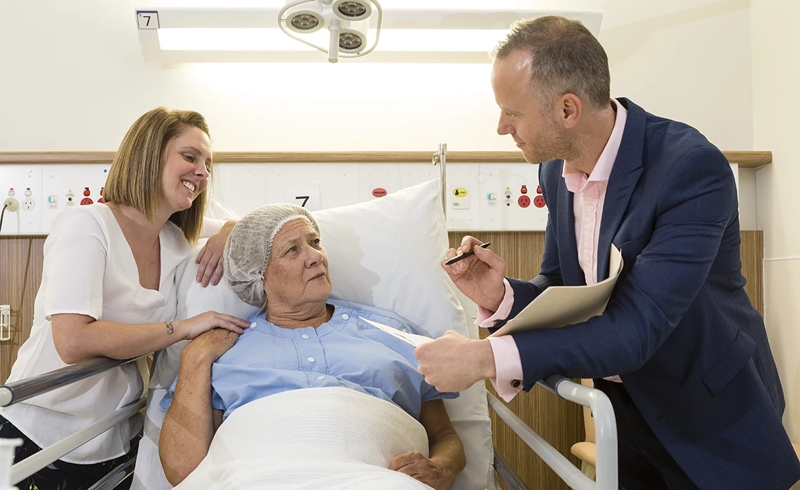 A patient in a hospital bed engaging in discussion with an anaesthetist in a suit, while a supportive family member sits beside them, smiling.