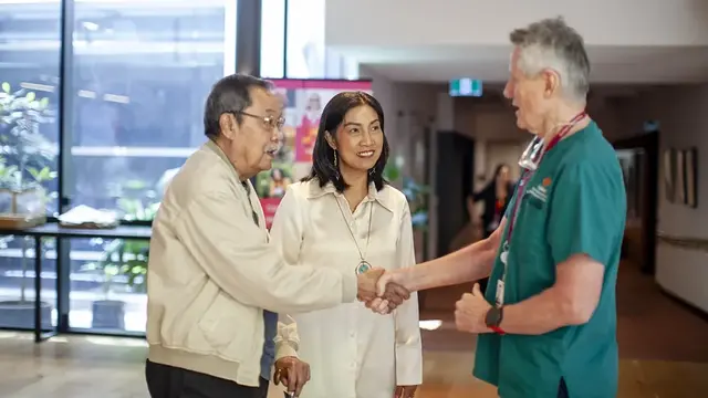 A friendly interaction in a medical setting, where an older man shakes hands with an anaesthetist while a caregiver smiles beside them