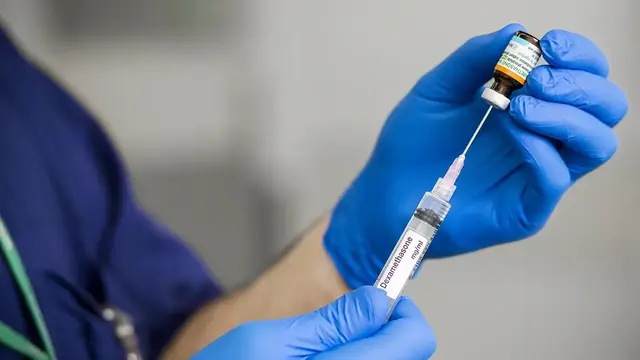 An anaesthetist in blue gloves prepares a syringe with Dexamethasone, drawing the medication from a vial in a clinical setting.