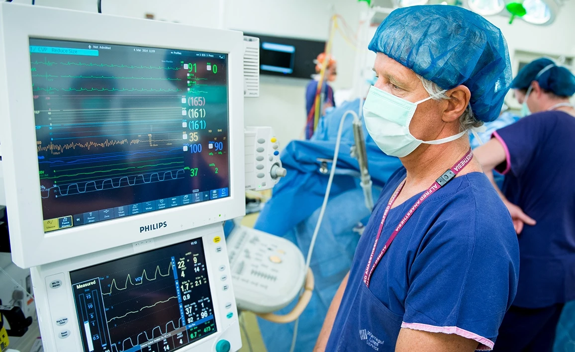 A masked and capped anaesthetist monitors vital signs on a screen in a busy operating room, with surgical staff working in the background.