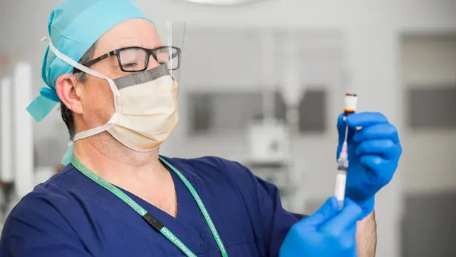 Doctor in scrubs and face mask with glasses drawing up needle of medicine
