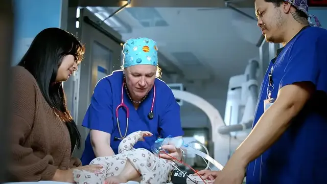 A team of healthcare professionals in scrubs and a caregiver attend to an infant on a hospital bed, providing respiratory support in a clinical setting. 