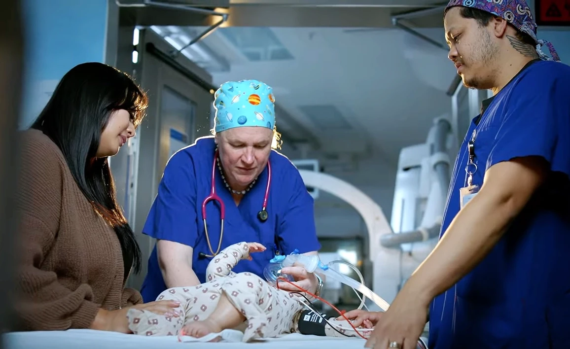 A team of healthcare professionals in scrubs and a caregiver attend to an infant on a hospital bed, providing respiratory support in a clinical setting. 