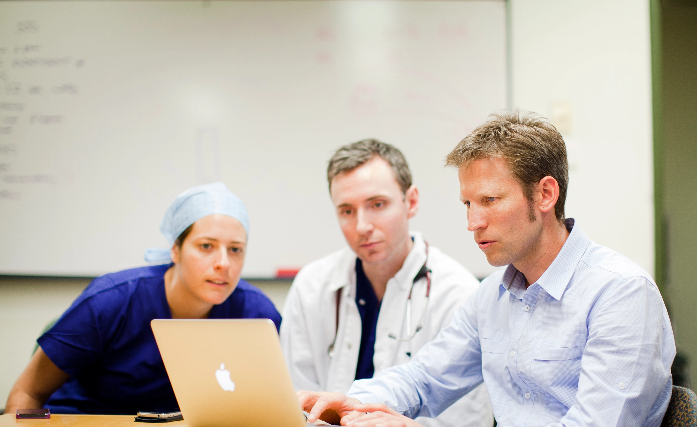 Three people looking at a mac laptop. One is wearing blue scrubs and hat, one wearing a white lab coat and the other a blue shirt