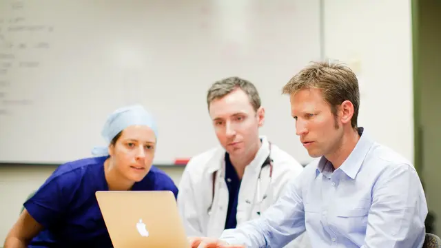Three people looking at a mac laptop. One is wearing blue scrubs and hat, one wearing a white lab coat and the other a blue shirt