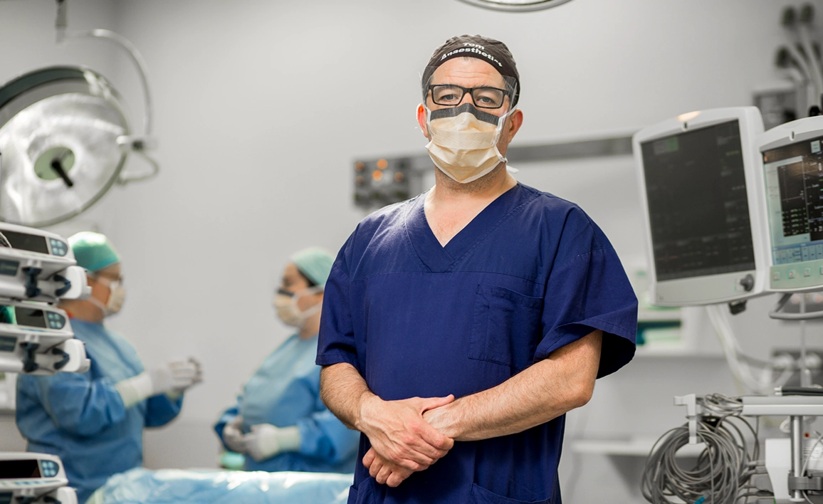 Male anaesthetist facing camera in operating theatre, wearing scrubs. Other doctor's chat in the background. 