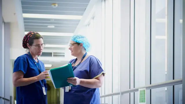 Two people in blue scrubs talking in a hospital corridor. One is holding a folder of papers