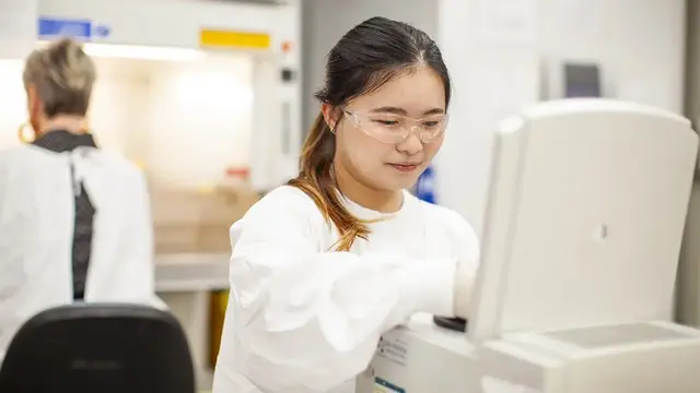 A scientist in a lab coat, safety glasses, and gloves operates laboratory equipment, while another person works near a fume hood in a research lab.