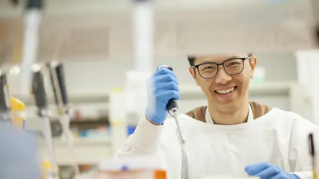 A smiling scientist in a lab, wearing safety glasses, gloves, and a white coat. He holds a pipette, working with samples. The background features lab equipment, including pipettes and shelves with supplies, emphasising a research setting.