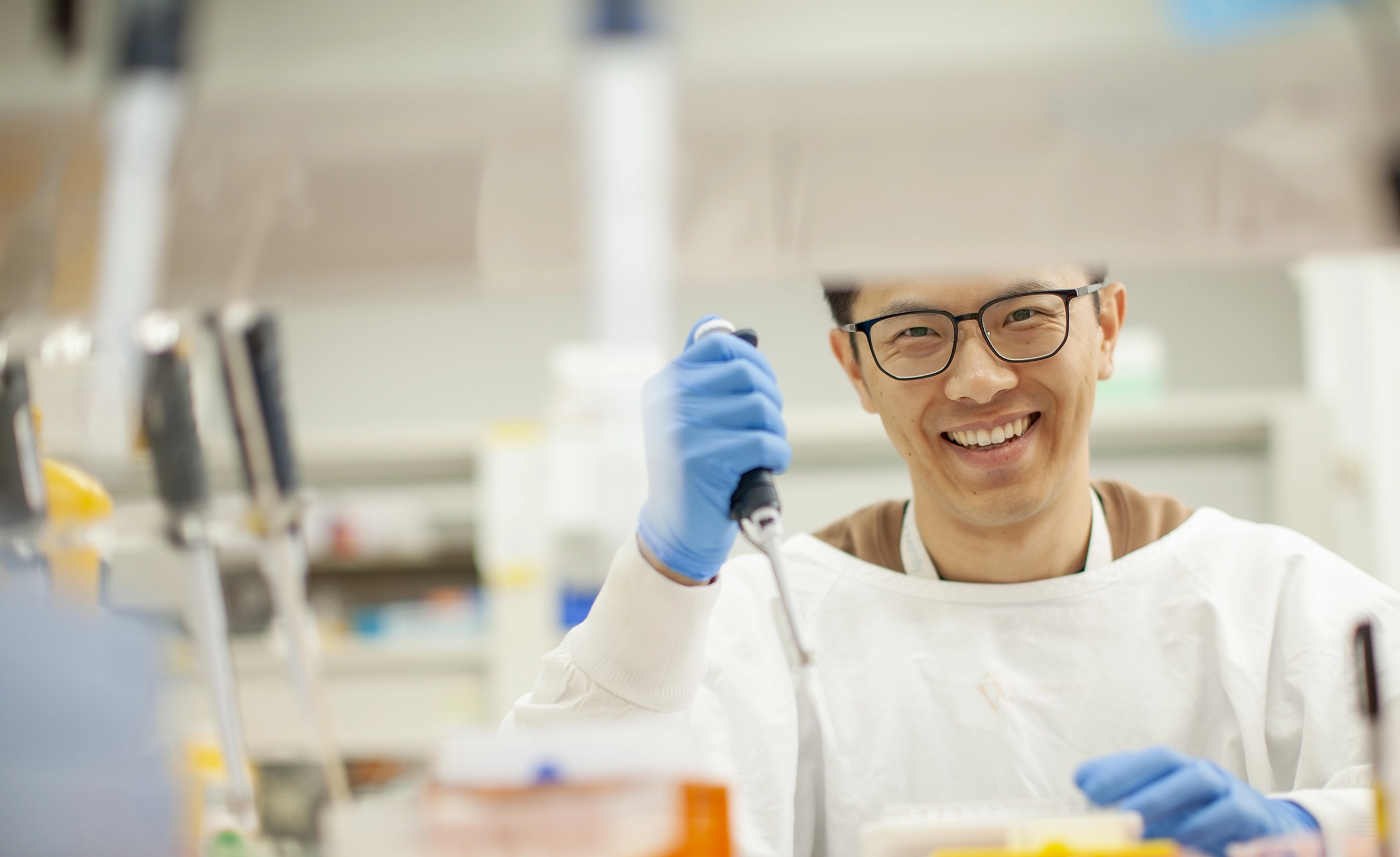 A smiling scientist in a lab, wearing safety glasses, gloves, and a white coat. He holds a pipette, working with samples. The background features lab equipment, including pipettes and shelves with supplies, emphasising a research setting.