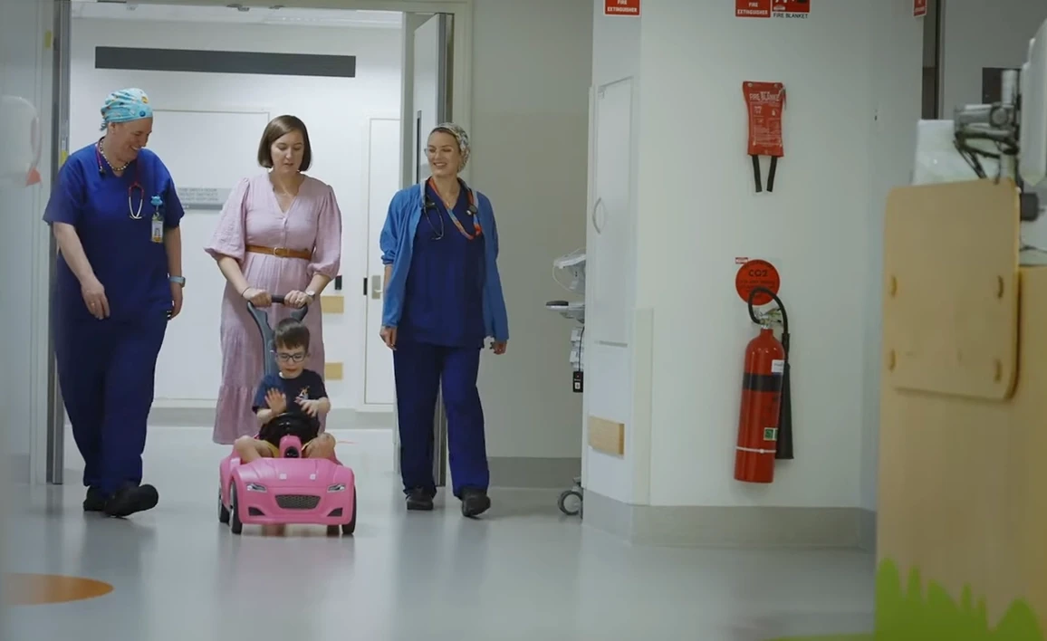 A young child rides a pink toy car down a hospital hallway, accompanied by a caregiver and two anaesthetists in scrubs, creating a supportive and comforting environment.