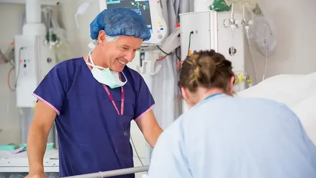 A smiling anaesthetist in scrubs and a surgical cap interacts with a colleague at a patient’s bedside in a hospital intensive care unit.