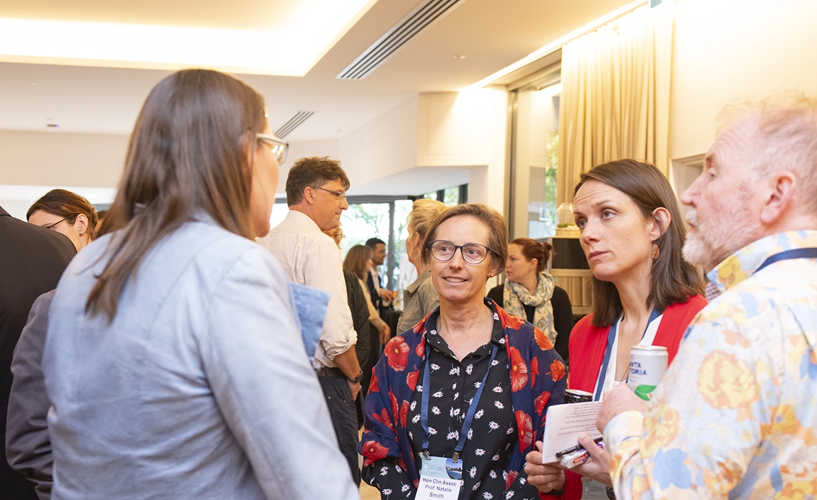 A professional networking event where four individuals engage in conversation, with other attendees mingling in the background.