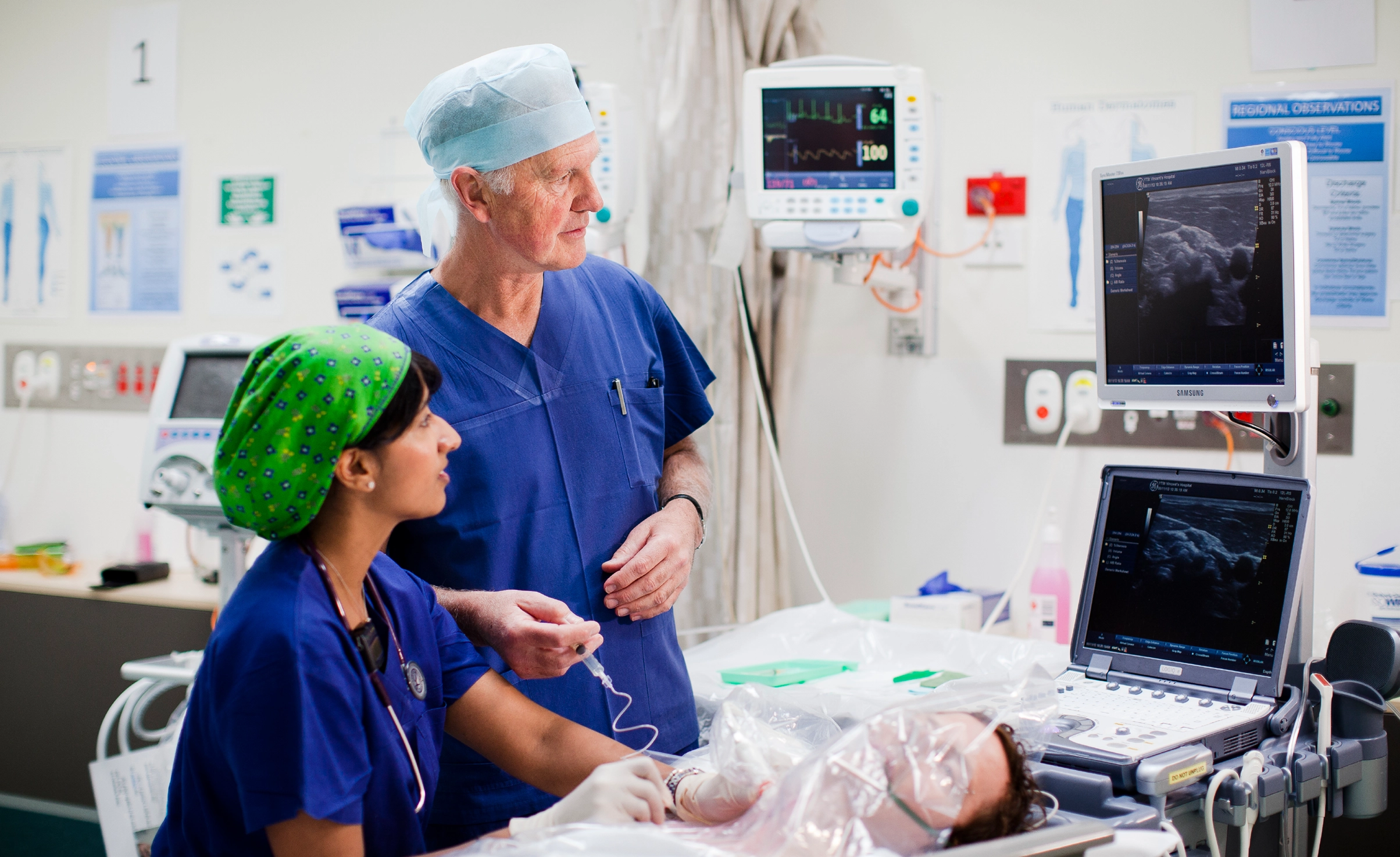 Male and female anaesthetists conduct a review at the bedside of an unconcious patient in a hospital setting. 