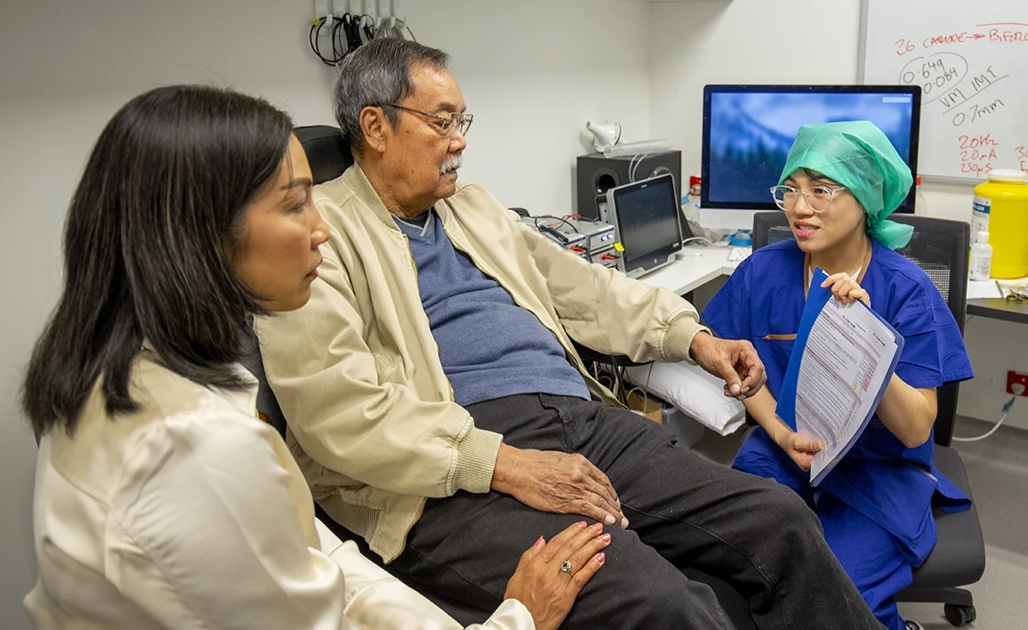 A research coordinator in blue scrubs discusses a medical document with a patient and a companion in a clinical setting, surrounded by medical equipment.