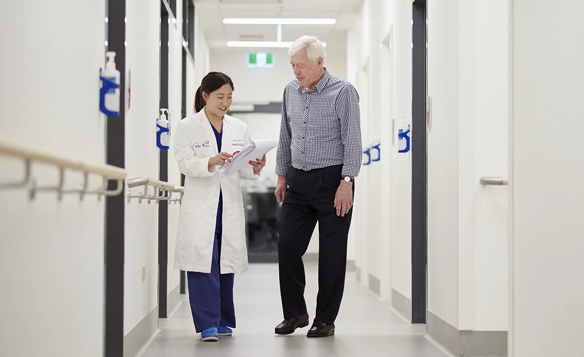 An anaesthetist in a white coat walks alongside an elderly patient in a medical facility, discussing paperwork in a bright, modern hallway.