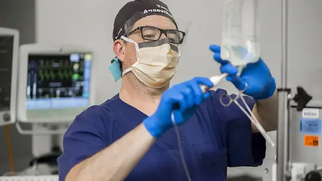 An anaesthetist in PPE prepares an IV infusion in an operating room, with medical monitors displaying vital signs in the background.