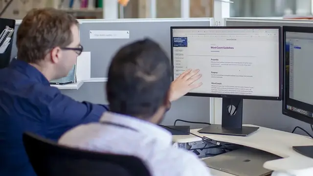 Two professionals at a workstation, discussing research information displayed on a computer screen in an office setting.