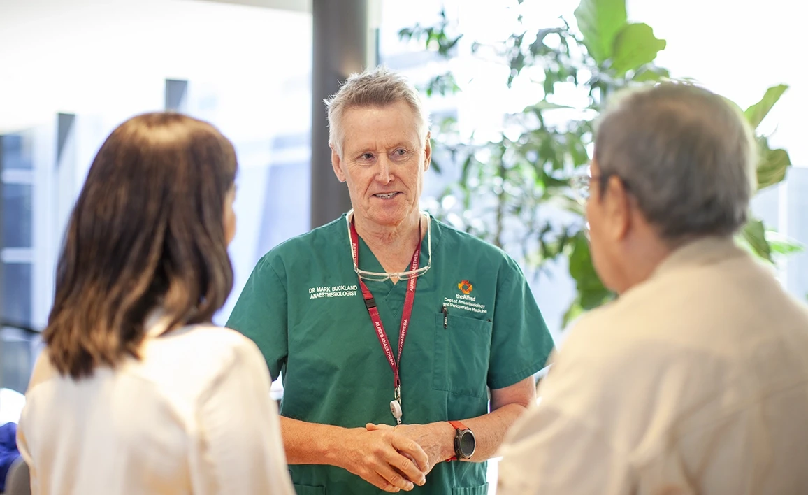 An anaesthetist in green scrubs, wearing a lanyard, engages in conversation with two individuals in a well-lit setting with greenery in the background.