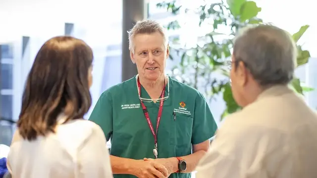 An anaesthetist in green scrubs, wearing a lanyard, engages in conversation with two individuals in a well-lit setting with greenery in the background.