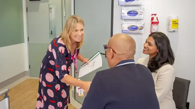 The image shows a friendly interaction in hospital setting. A staff member is warmly greeting a seated man with a handshake, while another family member looks on smiling.